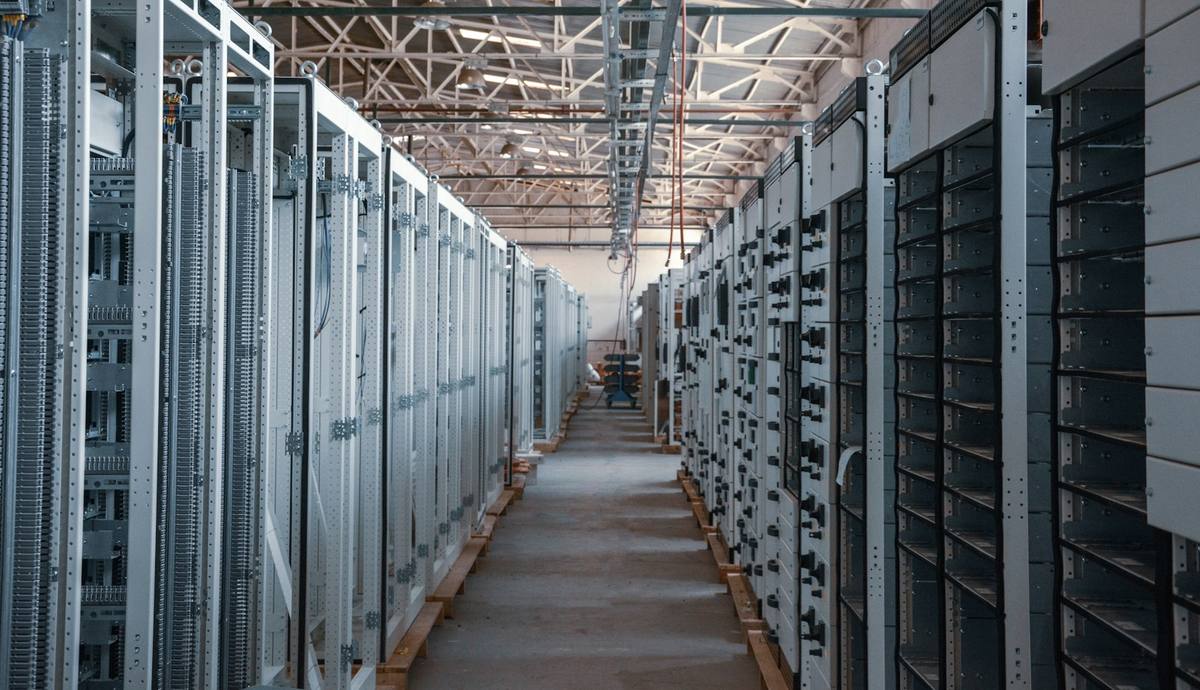 Aisle between rows of equipment cabinets under a steel roof truss in an industrial hall